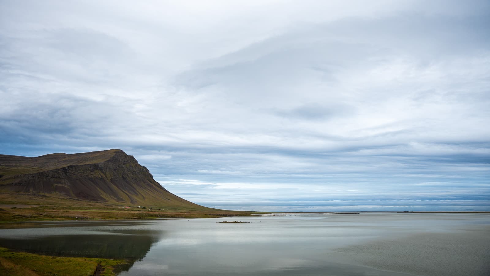 A flat-topped mountain reflected in the still shallows of an Icelandic fjord beneath an overcast sky