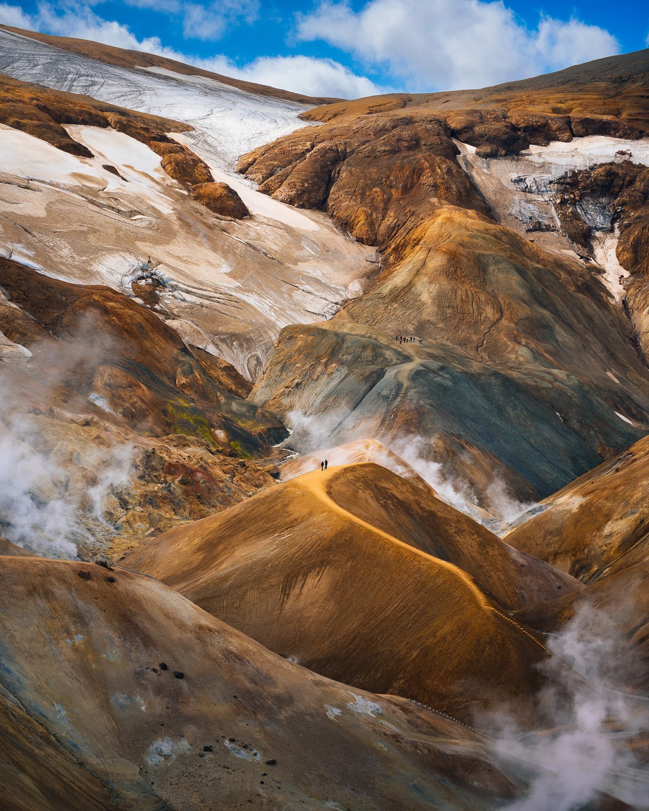 Snow and steam among ochre geothermal ridges in Iceland, with tiny hikers on the slopes
