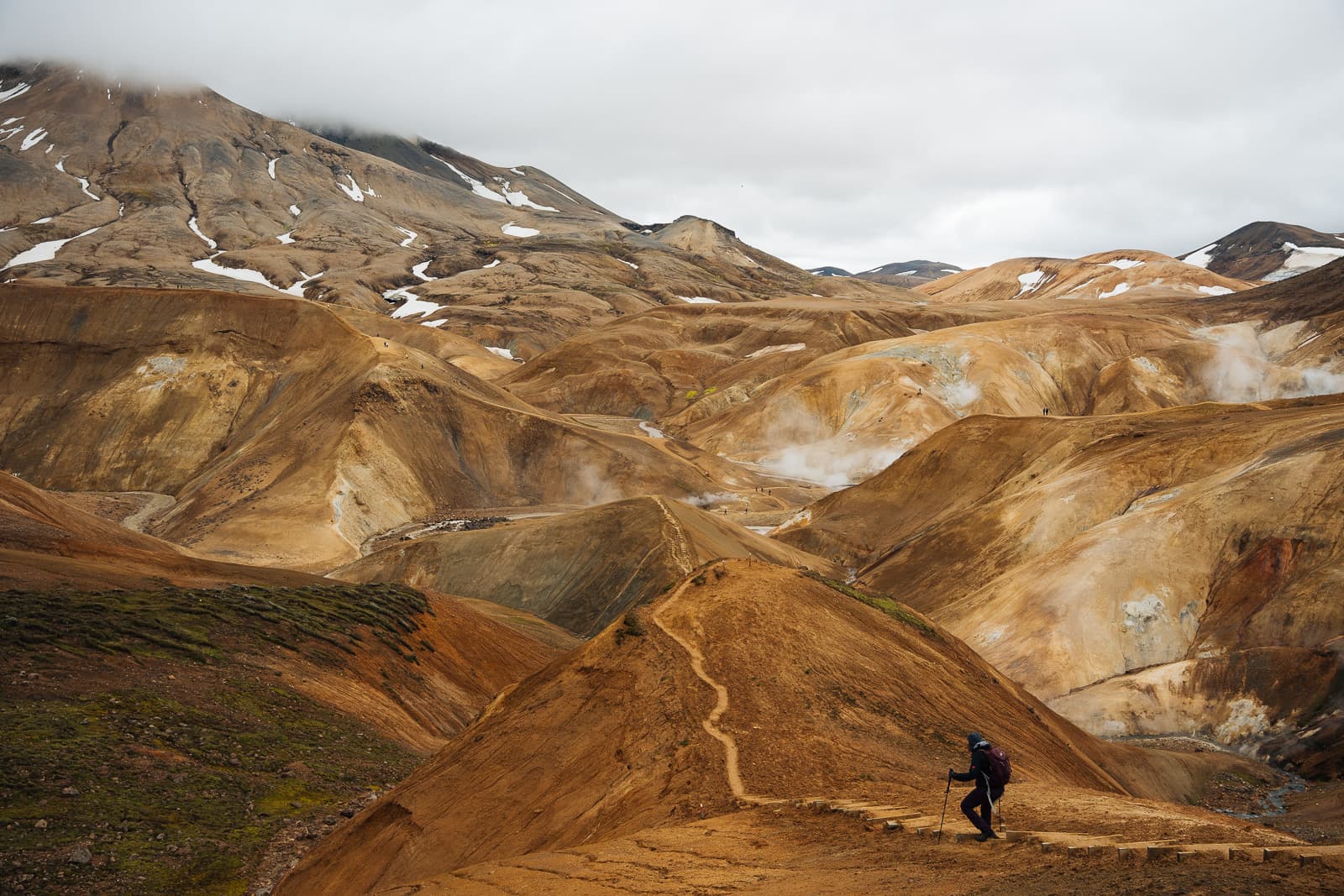 A hiker crossing an ochre ridge beneath snow-streaked mountains in the Icelandic highlands